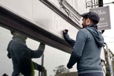 A man on a ladder fixing a painted name sign onto a bracket on a shopfront.