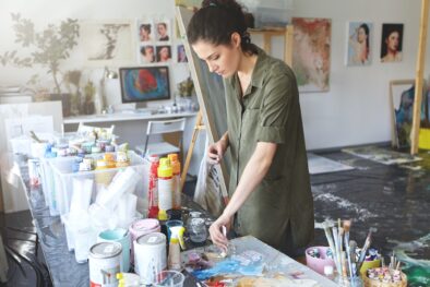 Busy female painter taking oil colors while standing near table with oils, working in art studio, go