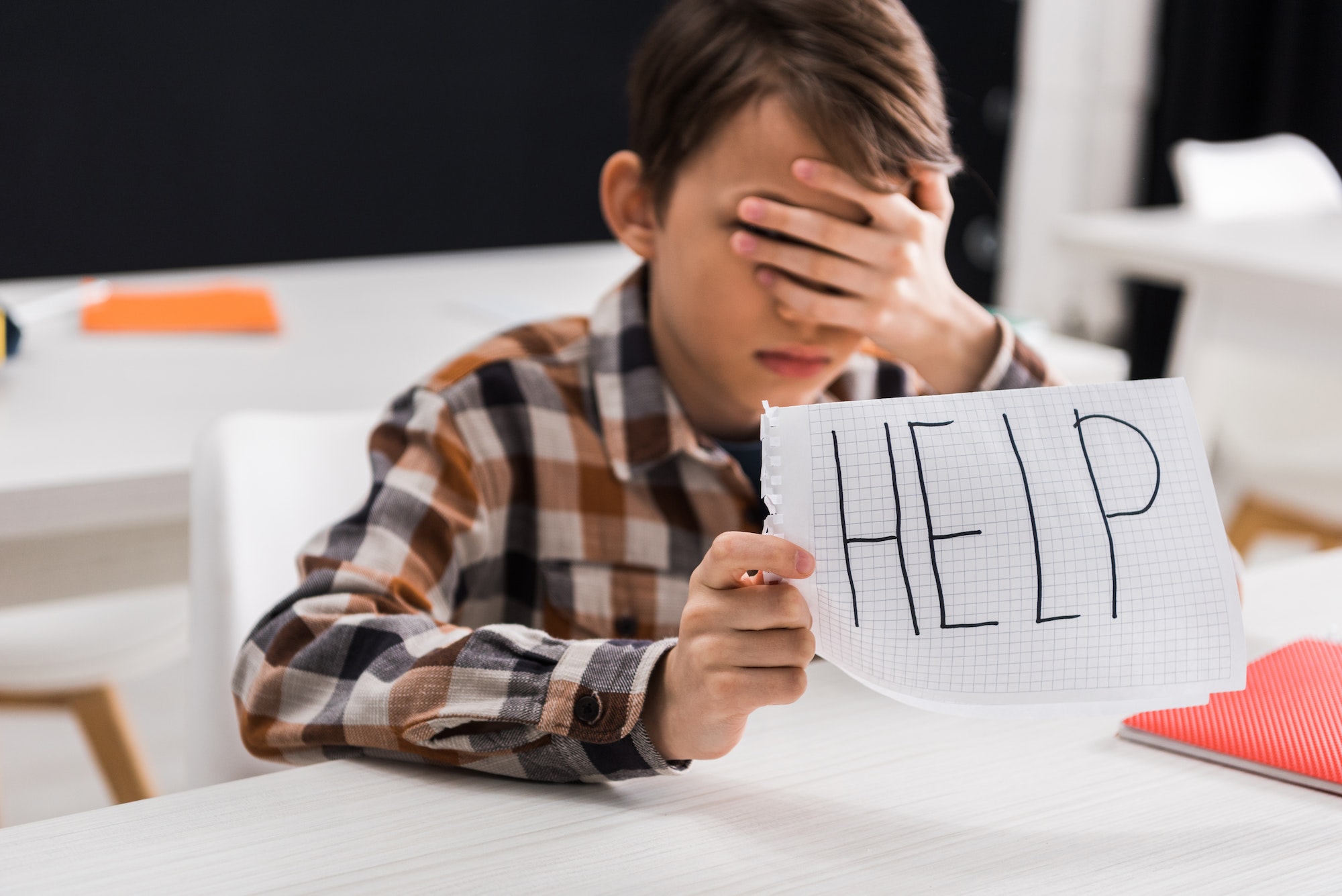 selective focus of upset schoolboy holding paper with help lettering and covering eyes, bullying