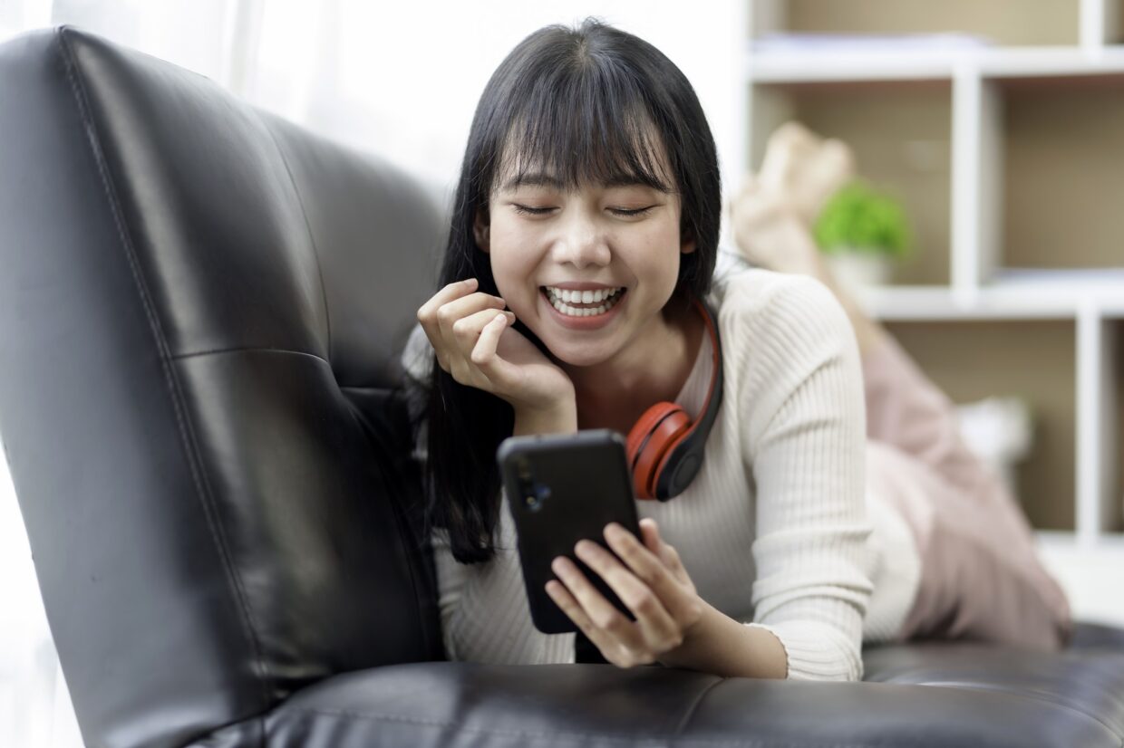 Cute beautiful young Asian woman lying on the sofa relaxing on the sofa playing social media wearing