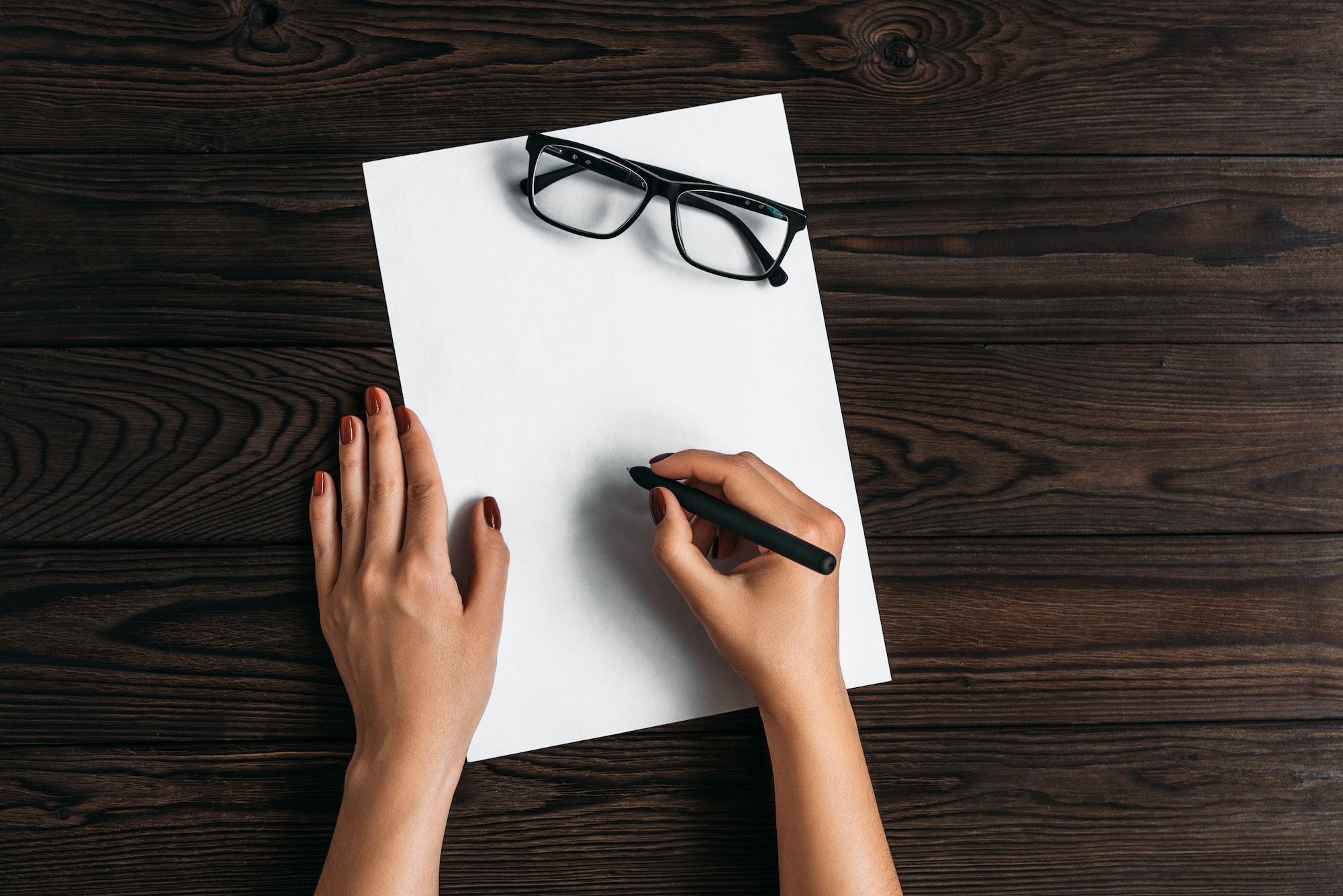 Write a letter on a white sheet. Hands, blank sheet, pen and glasses on wooden table